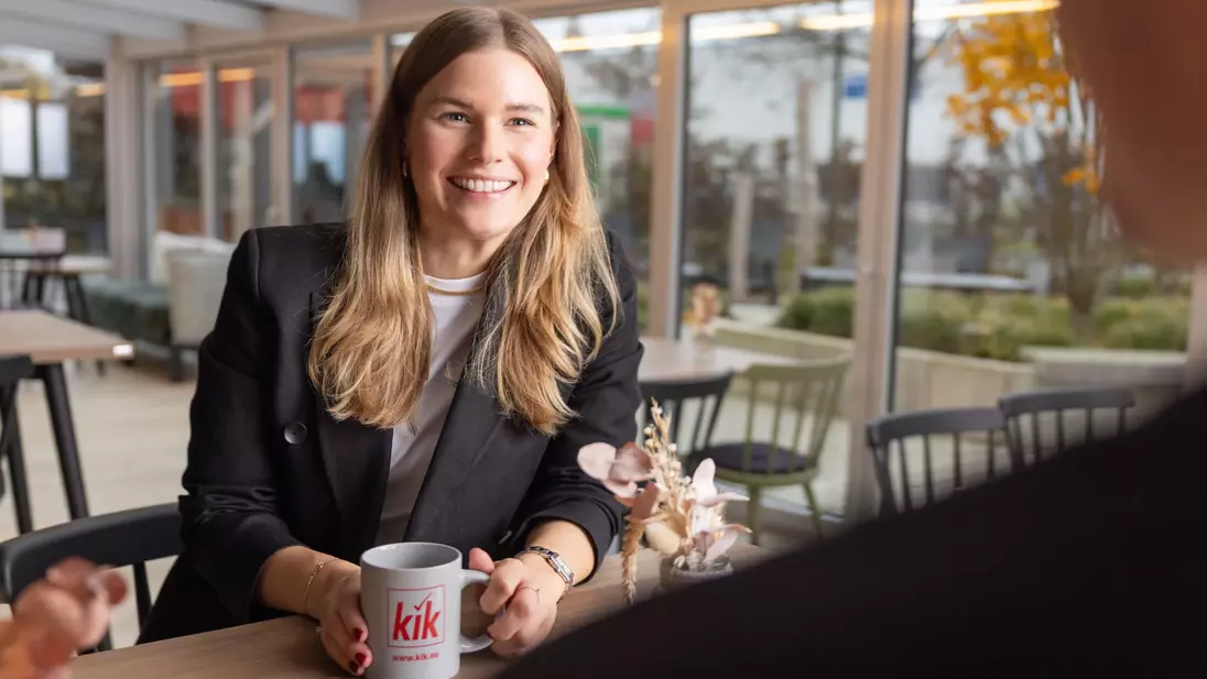 Een vrouw zit glimlachend aan een tafel, houdt een kopje met het ‘kik’-logo in haar hand en voert een gesprek.