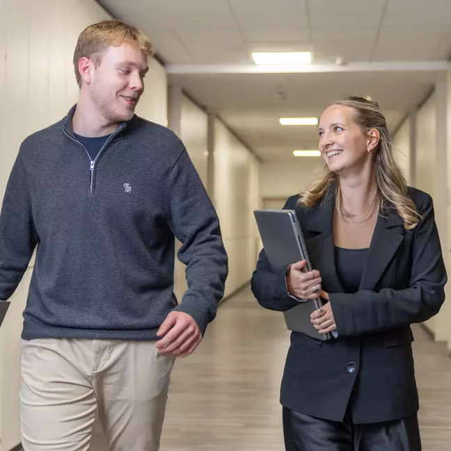 Twee medewerkers lopen door de gang van KiK met een laptop in de hand en glimlachen naar elkaar
