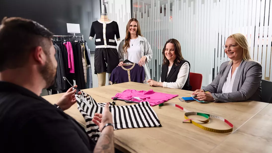 Vier mensen zitten en staan in een vergaderruimte aan een tafel met kledingstukken, stofstalen en een meetlint, en bespreken een modecollectie.