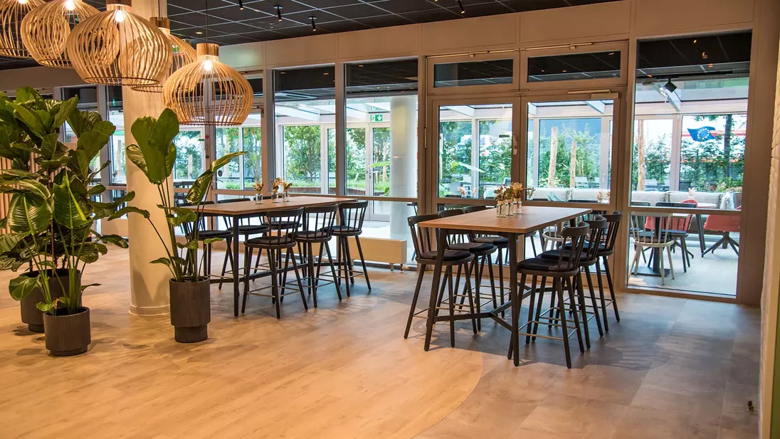 Interieur van een moderne kantine met tafels, hoge zitplaatsen, grote ramen en planten.