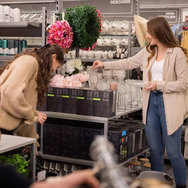 Twee medewerksters sorteren decoratieartikelen in de schappen van een winkel in woonaccessoires.