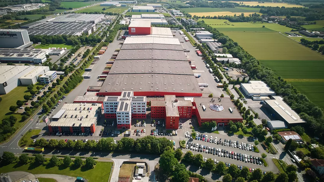 Luchtfoto van het hoofdkantoor met lange haldaken, rode gebouwdelen, parkeerplaatsen en omliggende velden.
