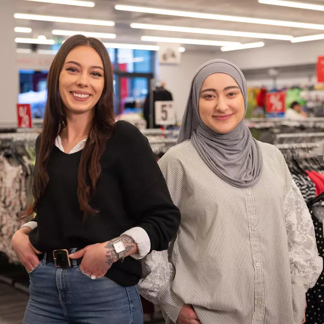 Twee medewerkers staan glimlachend voor kledingrekken in een KiK-filiaal
