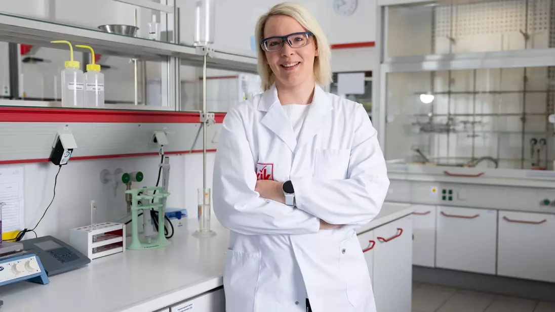 Een laboratoriummedewerkster in een witte laboratoriumjas staat met haar armen over elkaar in een modern laboratorium; ze draagt een veiligheidsbril en glimlacht.