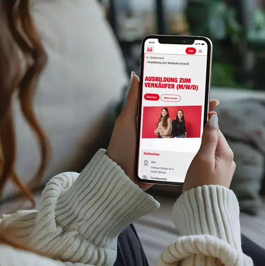 Eine Frau sitzt auf der Couch und schaut auf ihr Handydisplay, auf dem eine Stellenausschreibung von KiK angezeigt wird.