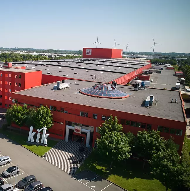 Een groot, rood bedrijfsgebouw met het logo „kik“ staat in een groen landschap.
