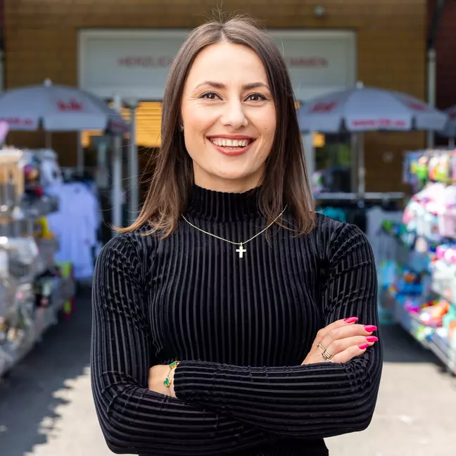 Een persoon staat buiten voor de winkel, tussen de schappen, en kijkt glimlachend naar de camera.