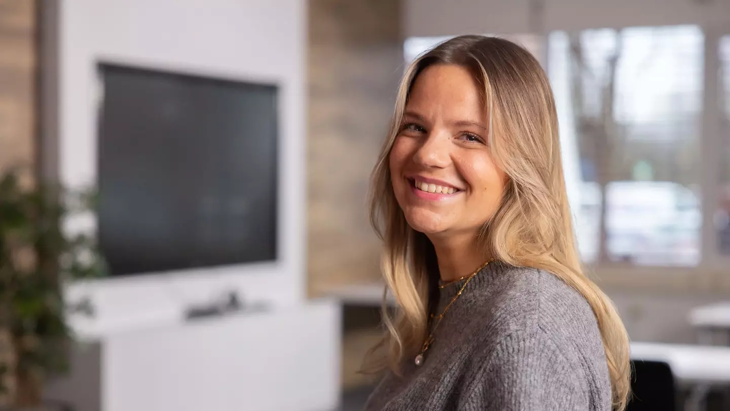 Een vrouw kijkt glimlachend in de camera; ze staat in een moderne ruimte