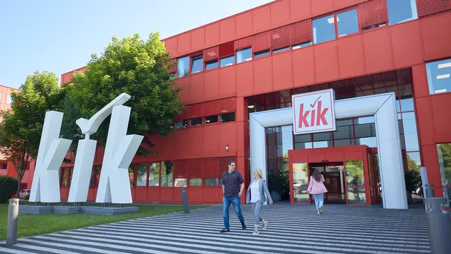 Het rode KiK-gebouw met een groot wit logo bij de ingang en verschillende mensen die het gebouw binnenlopen.